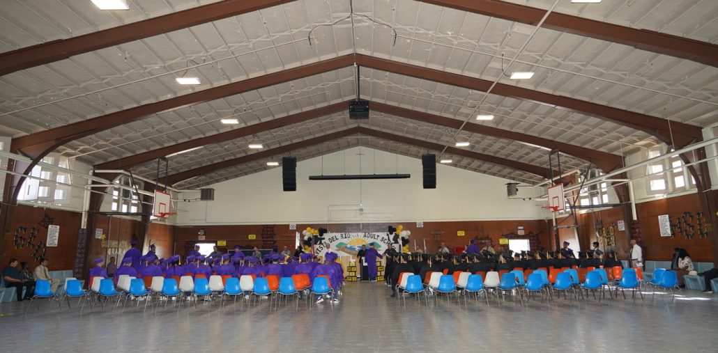 graduates sitting in a large room wearing their commencement cap and gowns and listneing to a speaker at the podium